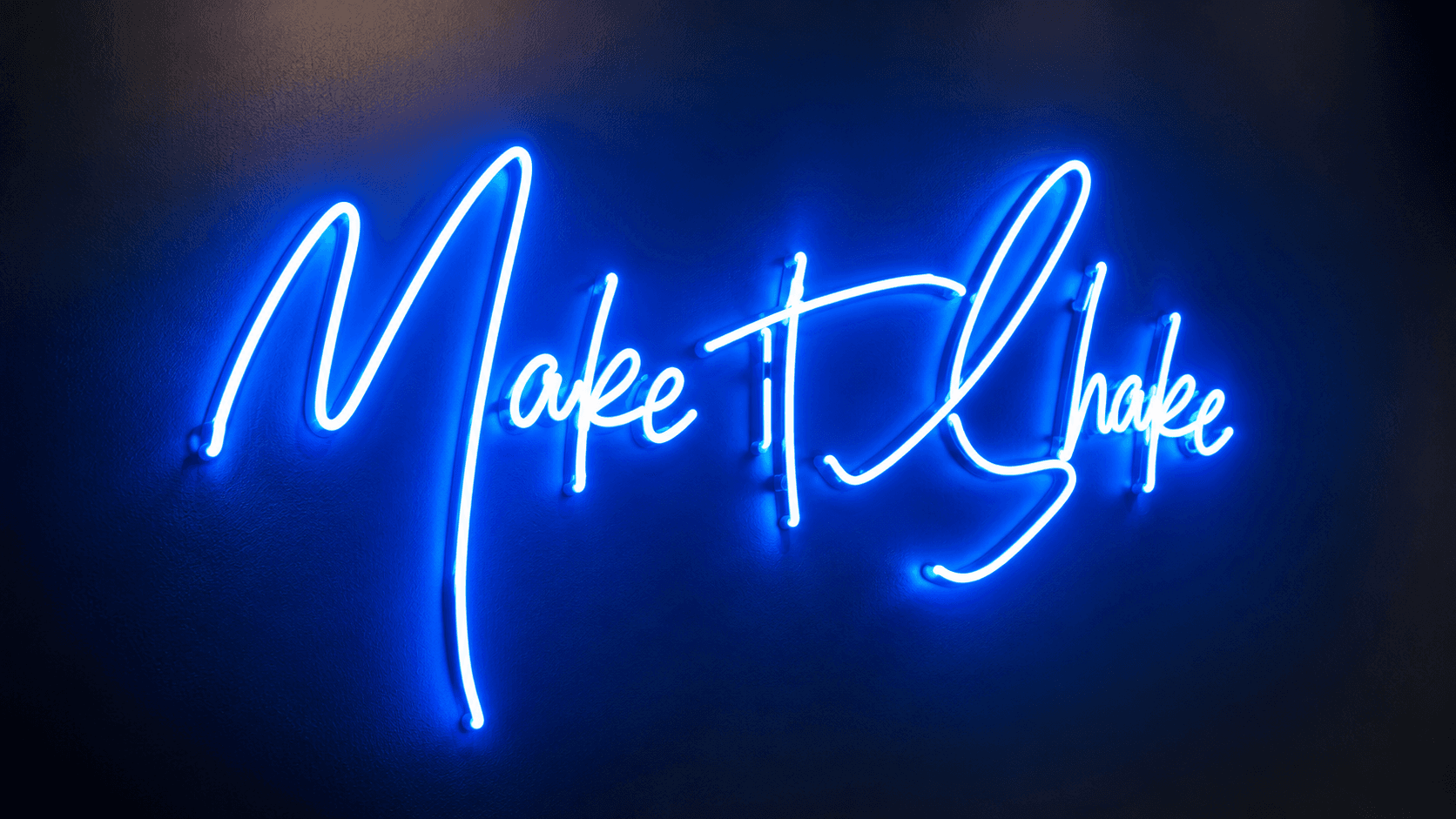 Blue neon “Make it Shake” script sign glowing on a dark studio wall