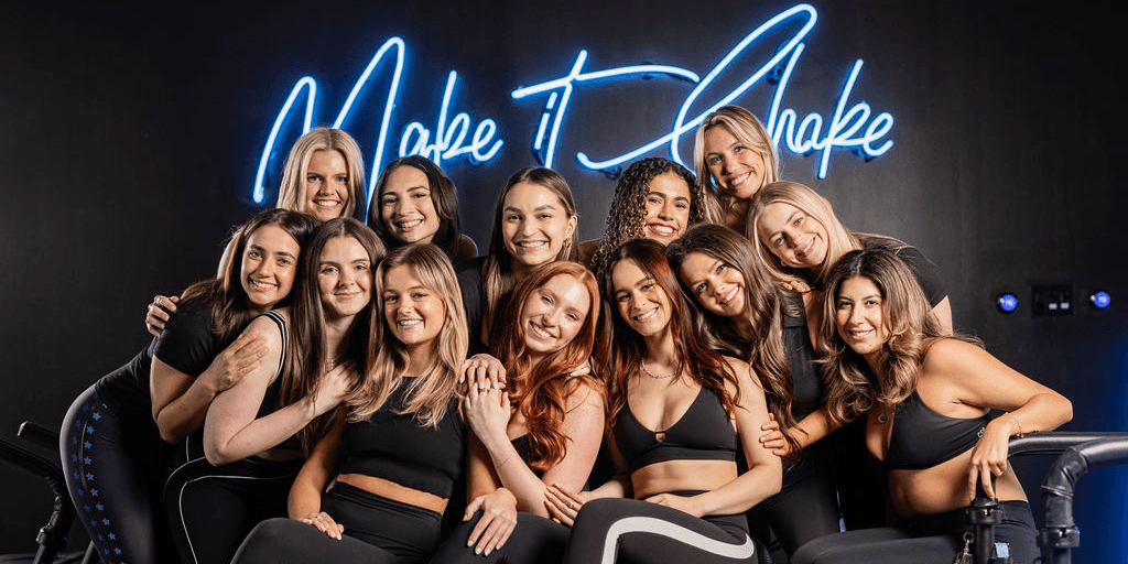 BE Fit instructors smiling together under the neon Make It Shake sign